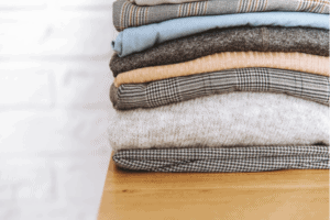 A pile of neatly folded thin sweaters that are neutral colors on a wood desk